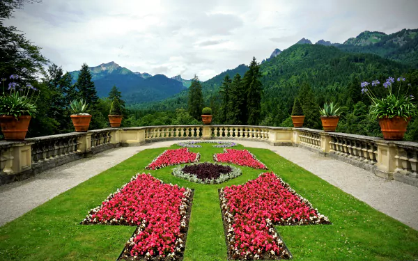 A vibrant garden with red flower beds arranged symmetrically in the courtyard of Linderhof Palace, with mountainous scenery in the background and a decorative fence lining the area.