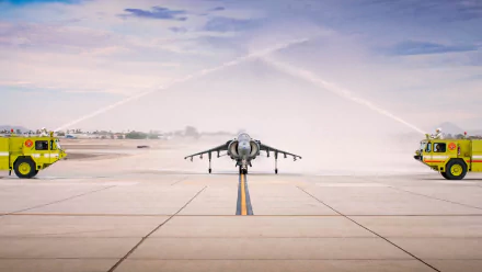 HD desktop wallpaper featuring a military jet fighter on a runway framed by two fire trucks spraying water arches over the aircraft.