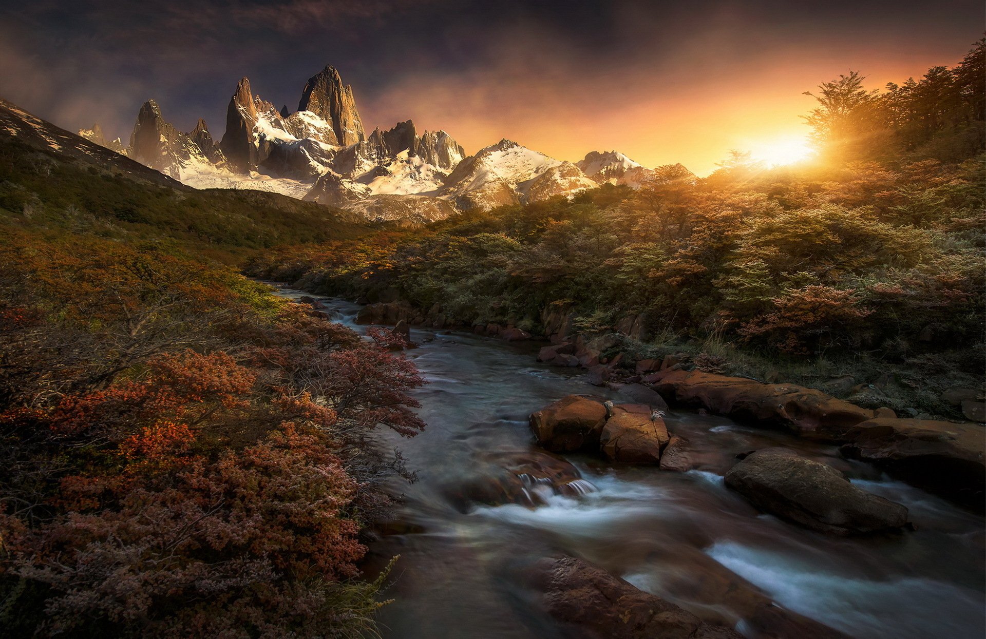 A stunning sunrise illuminates majestic mountain peaks, casting warm colors over a flowing stream, surrounded by vibrant nature—an HD wallpaper that captures the beauty of the outdoors.