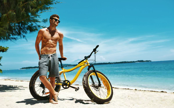 A shirtless man wearing sunglasses stands barefoot on sandy beach next to a yellow bicycle under a clear blue sky, captured in 4K Ultra HD.