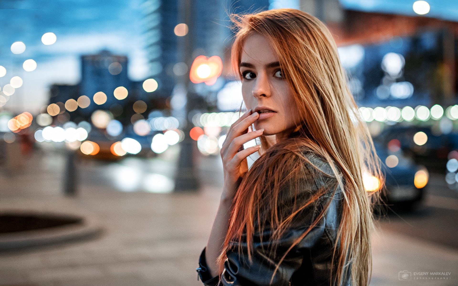 2K Quad HD PC desktop wallpaper: blonde woman with brown eyes in a leather jacket, shallow depth of field, city bokeh lights, looking over her shoulder.