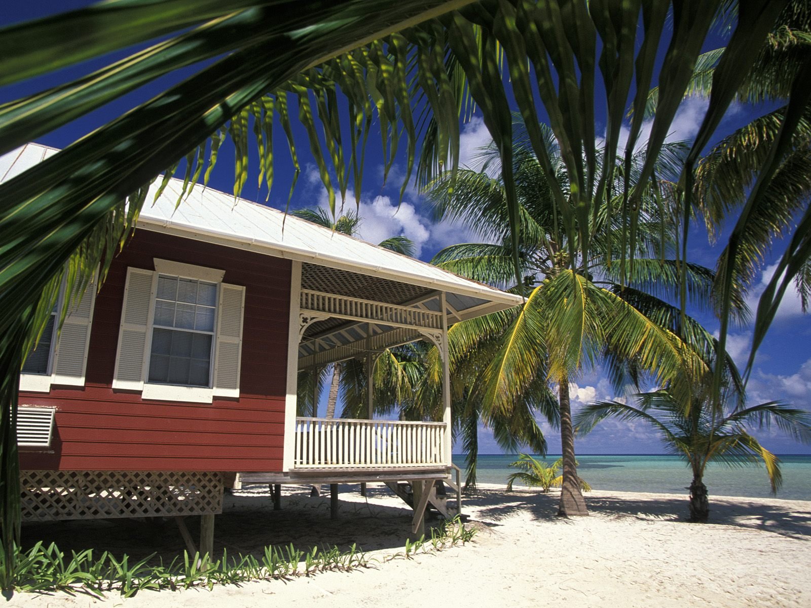 HD PC desktop wallpaper featuring a man-made house on a sandy beach, surrounded by palm trees under a bright blue sky.