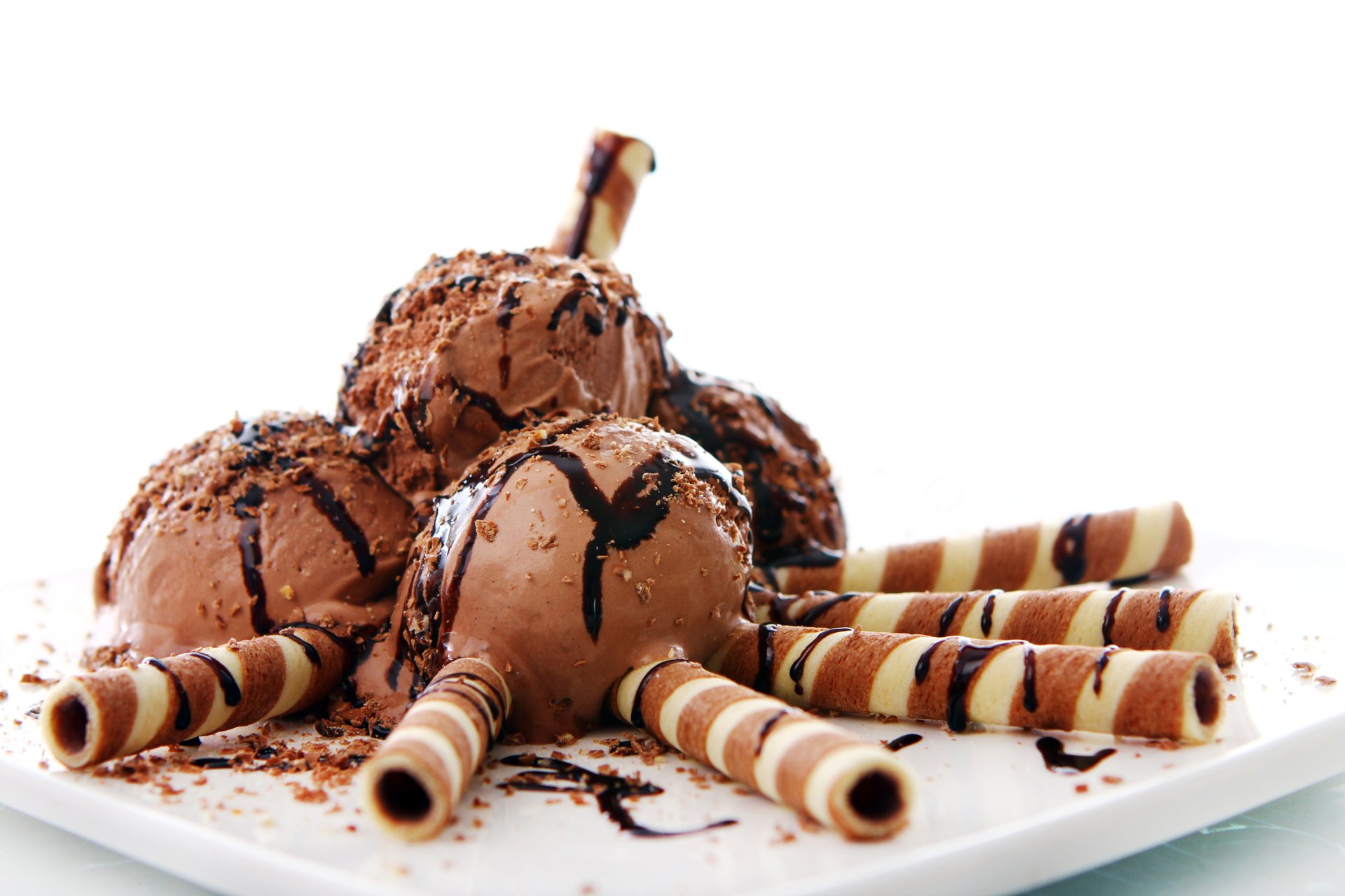 Close-up of chocolate ice cream scoops drizzled with chocolate sauce and surrounded by striped wafer rolls on a white plate, captured in stunning 4K Ultra HD.