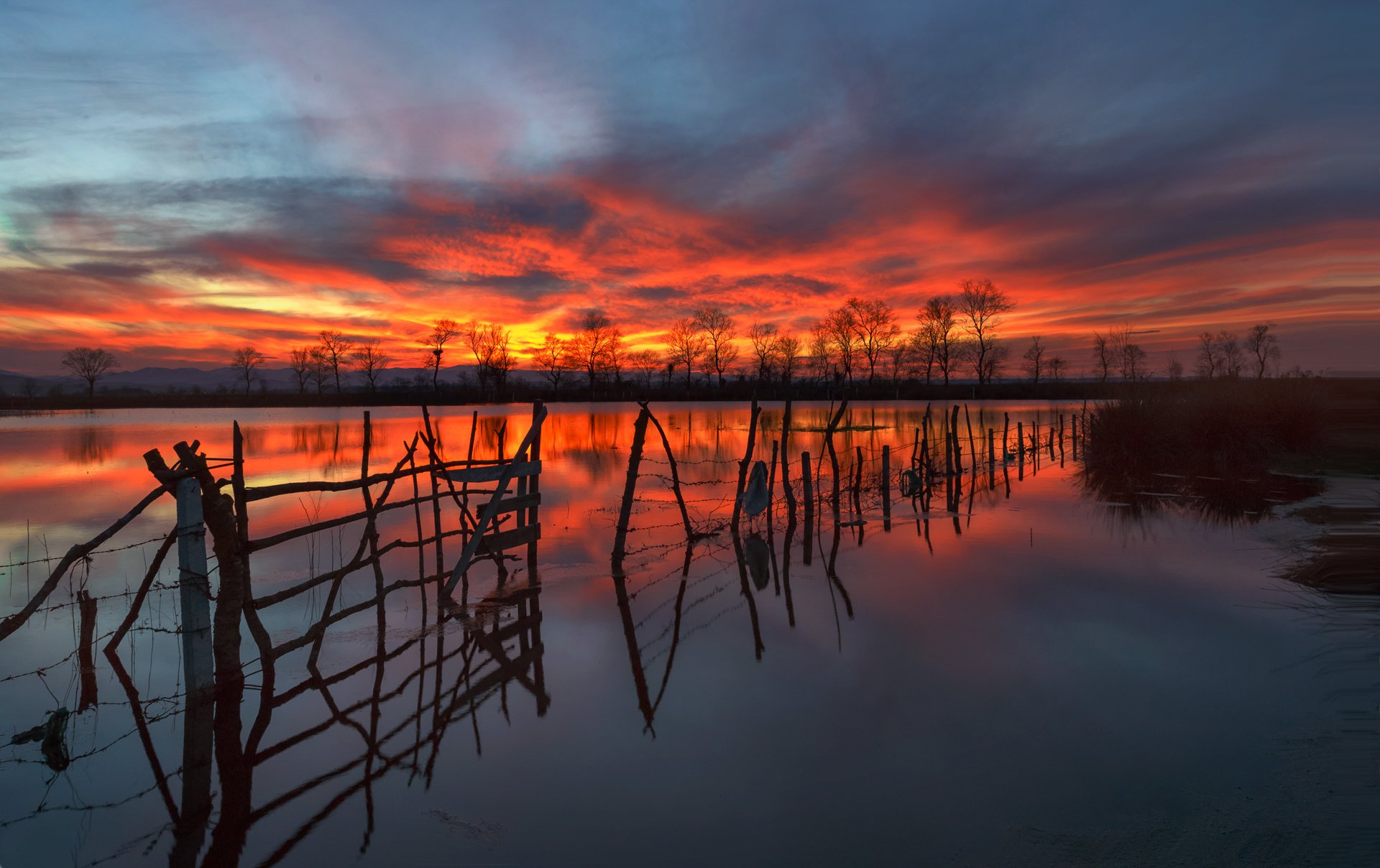 A stunning HD wallpaper featuring a serene lake with a rustic fence, captured at sunset. The vibrant sky and its reflection in the water create a picturesque natural scene.