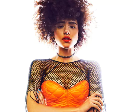 HD PC wallpaper of a brunette woman with curly hair, brown eyes and bold lipstick, wearing a choker and nose piercing, orange corset over a black fishnet top.