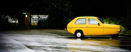 HD desktop wallpaper featuring a bright yellow Reliant Robin car parked on a wet road with a dark forest background.