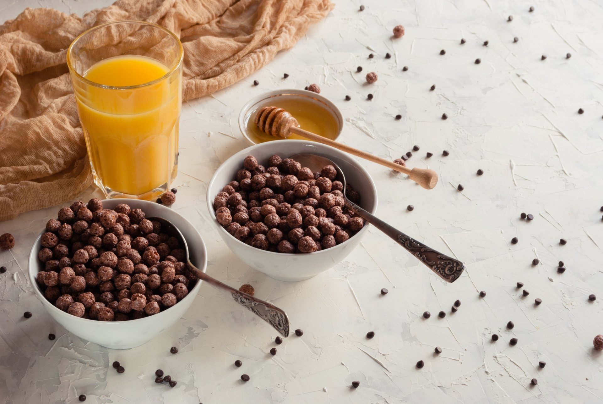 A 4K Ultra HD still life of a breakfast scene featuring two bowls of chocolate cereal, a glass of orange juice, and a honey dipper on a white textured surface.