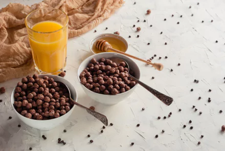 A 4K Ultra HD still life of a breakfast scene featuring two bowls of chocolate cereal, a glass of orange juice, and a honey dipper on a white textured surface.