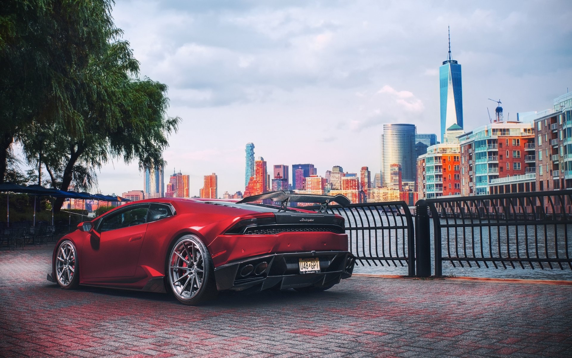 Red Lamborghini Huracan supercar parked by a waterfront with a city skyline in the background, captured as an HD PC desktop wallpaper and background.