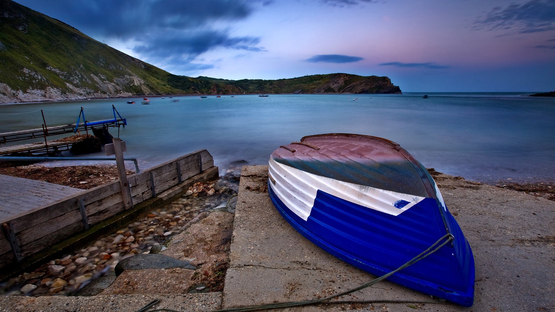 HD desktop wallpaper of a colorful boat resting on a concrete shore beside a rocky beach, with ocean waves, clouds, and a serene coastline under a vivid sky.