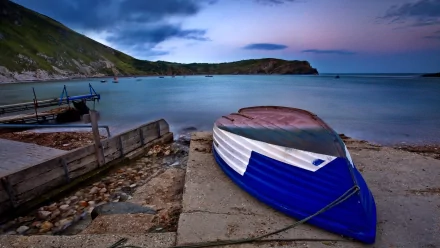 HD desktop wallpaper of a colorful boat resting on a concrete shore beside a rocky beach, with ocean waves, clouds, and a serene coastline under a vivid sky.