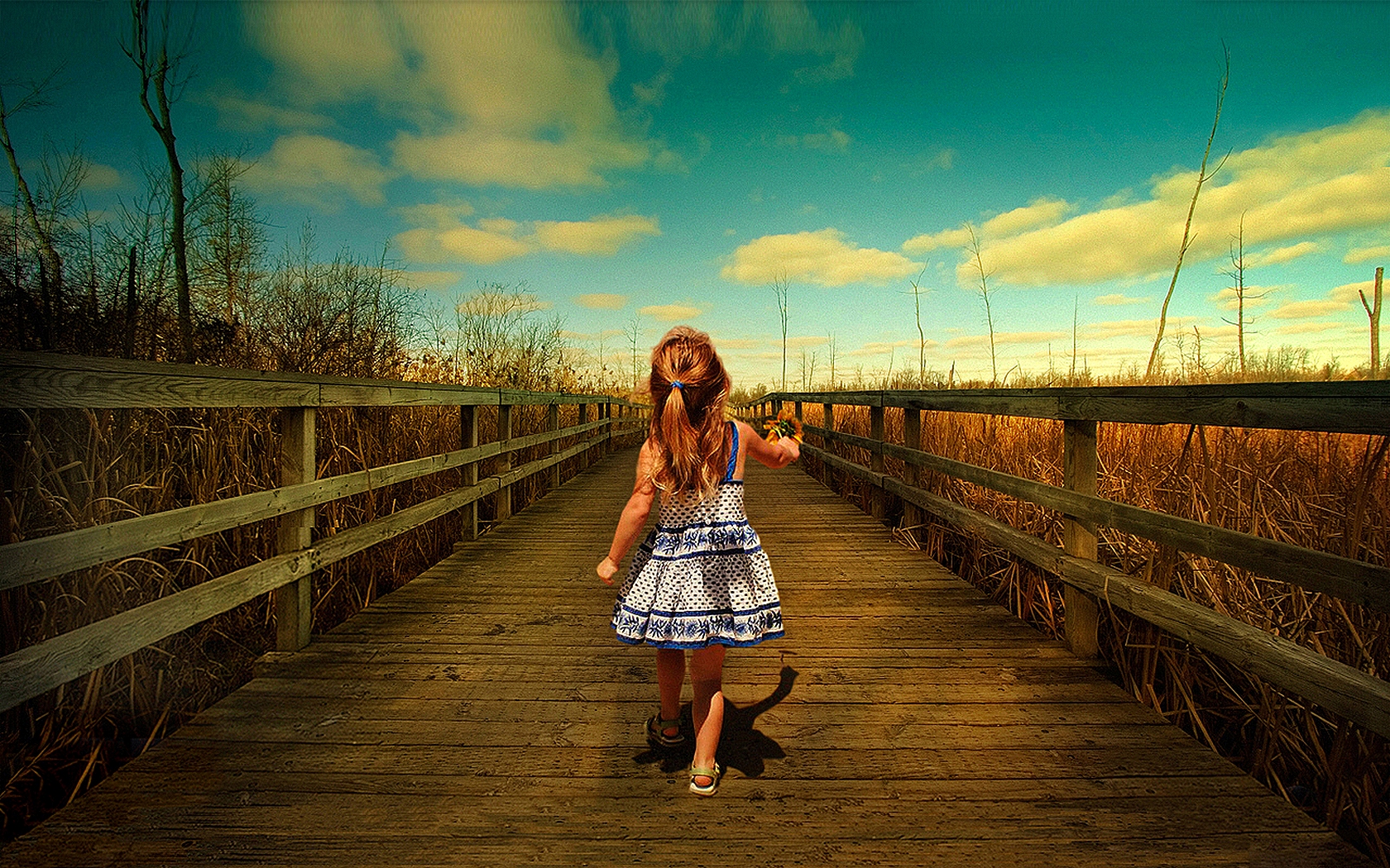 Cute little girl in a summer dress, standing on a bridge under the sun.