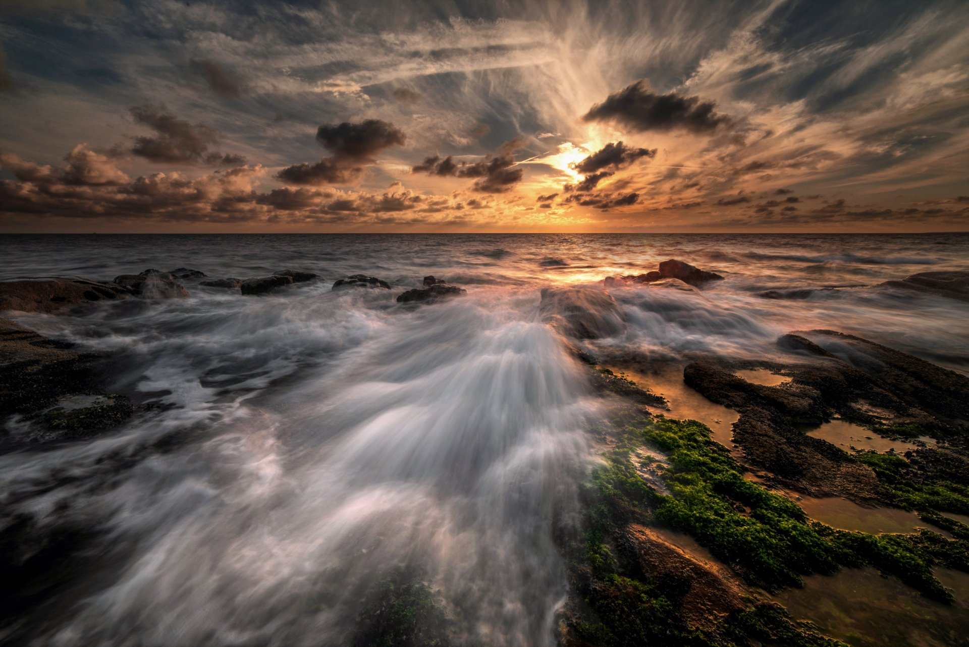 A breathtaking sunrise casts vibrant colors across the sky, reflecting on the ocean waves, with rocky shores and clouds enhancing the natural beauty in this HD desktop wallpaper.