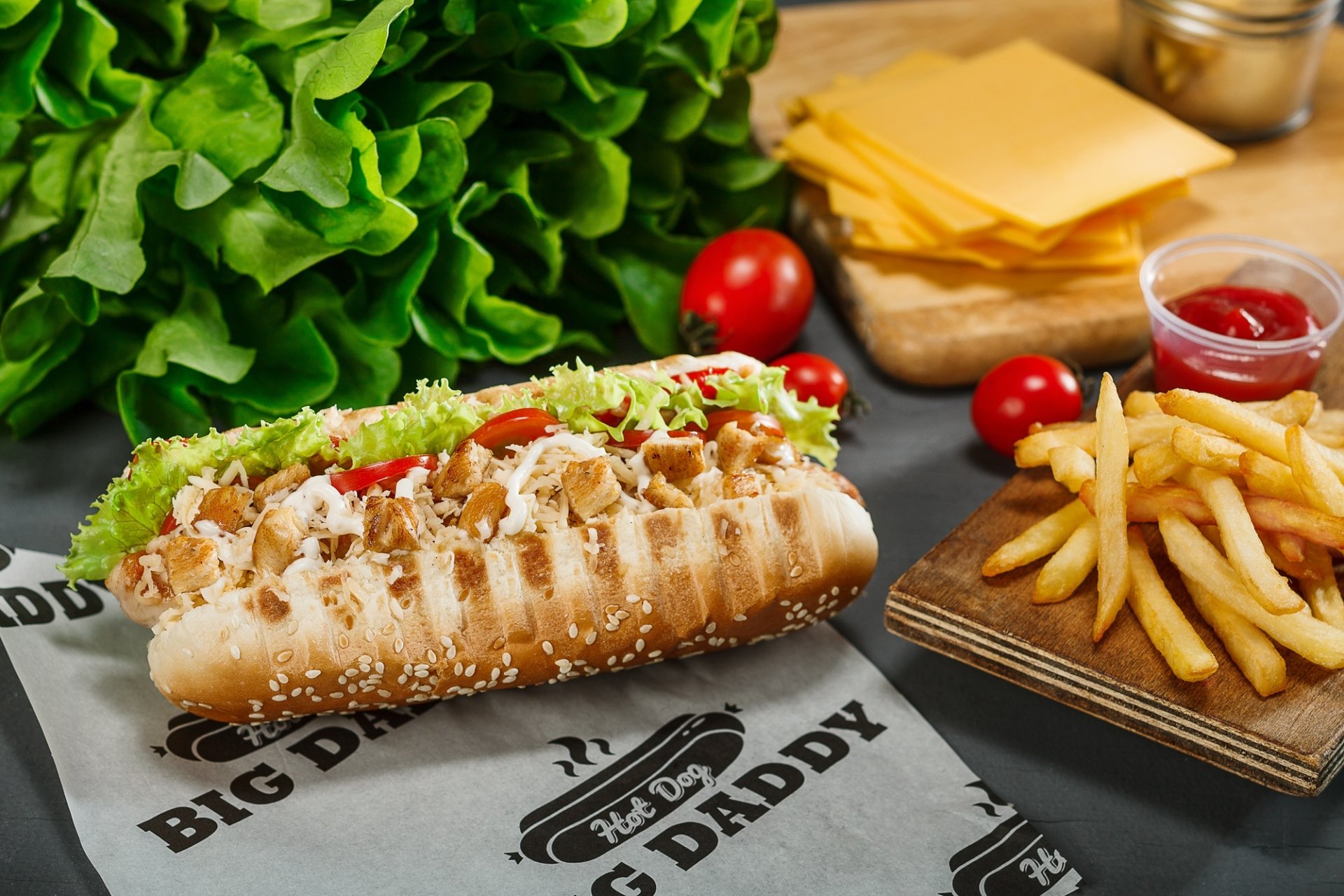 HD desktop wallpaper featuring a hot dog with cheese, lettuce, and tomato, accompanied by golden French fries and fresh ingredients in a vivid still life composition.