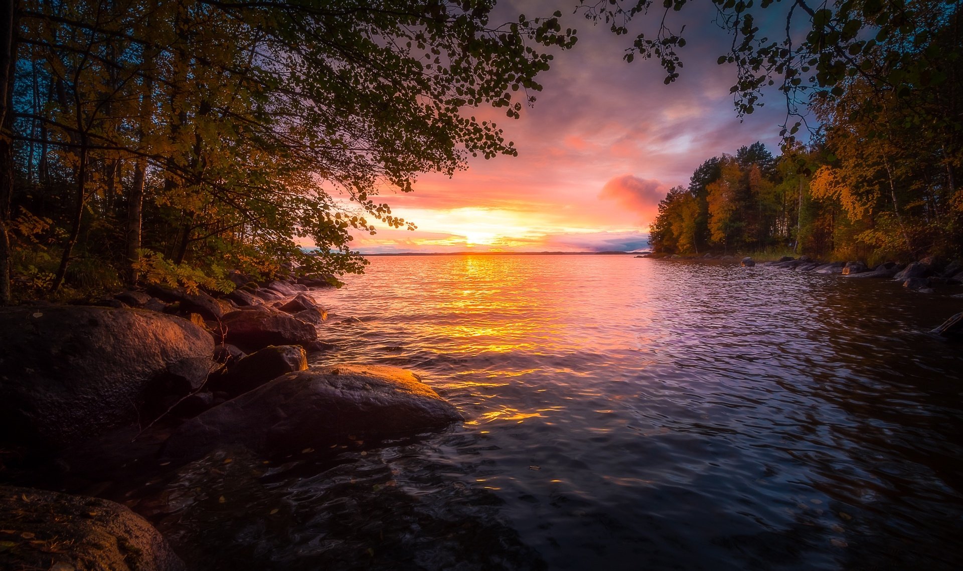 HD desktop wallpaper showing a tranquil lake at sunset, framed by lush trees and rocks. The sky is painted in vibrant hues of orange, pink, and purple, reflecting on the serene water.