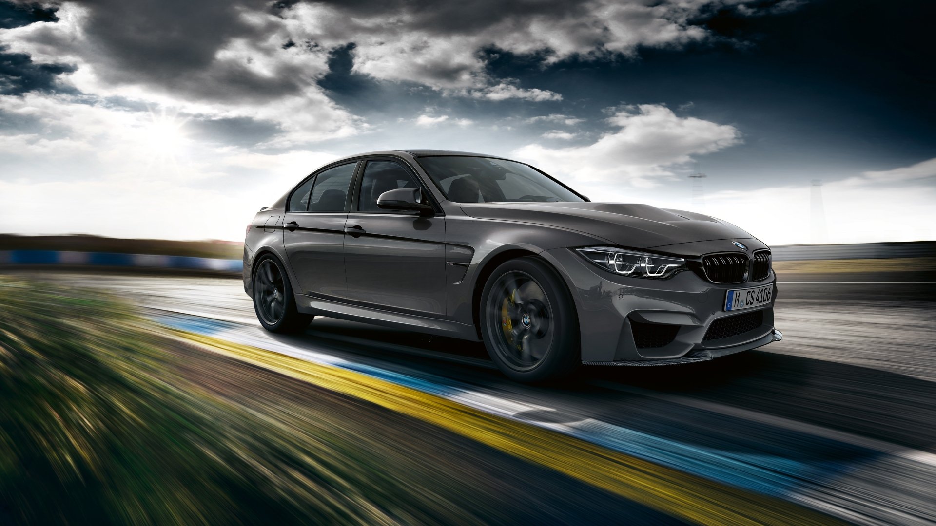 HD desktop wallpaper featuring a sleek silver BMW M3 car speeding on a racetrack under a dramatic cloudy sky.