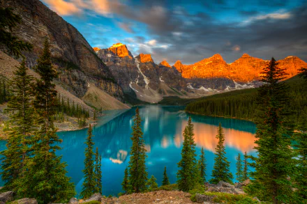 HD desktop wallpaper of Moraine Lake in Canada, featuring crystal-clear blue waters surrounded by lush forests, towering trees, and majestic mountains reflecting the golden hues of the setting sun.