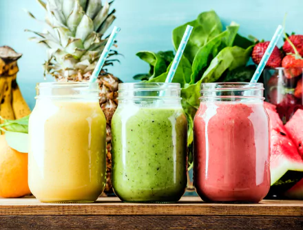 Colorful smoothie jars filled with yellow, green, and pink fruit drinks, surrounded by fresh fruits and greens on a wooden table. HD desktop wallpaper and background.