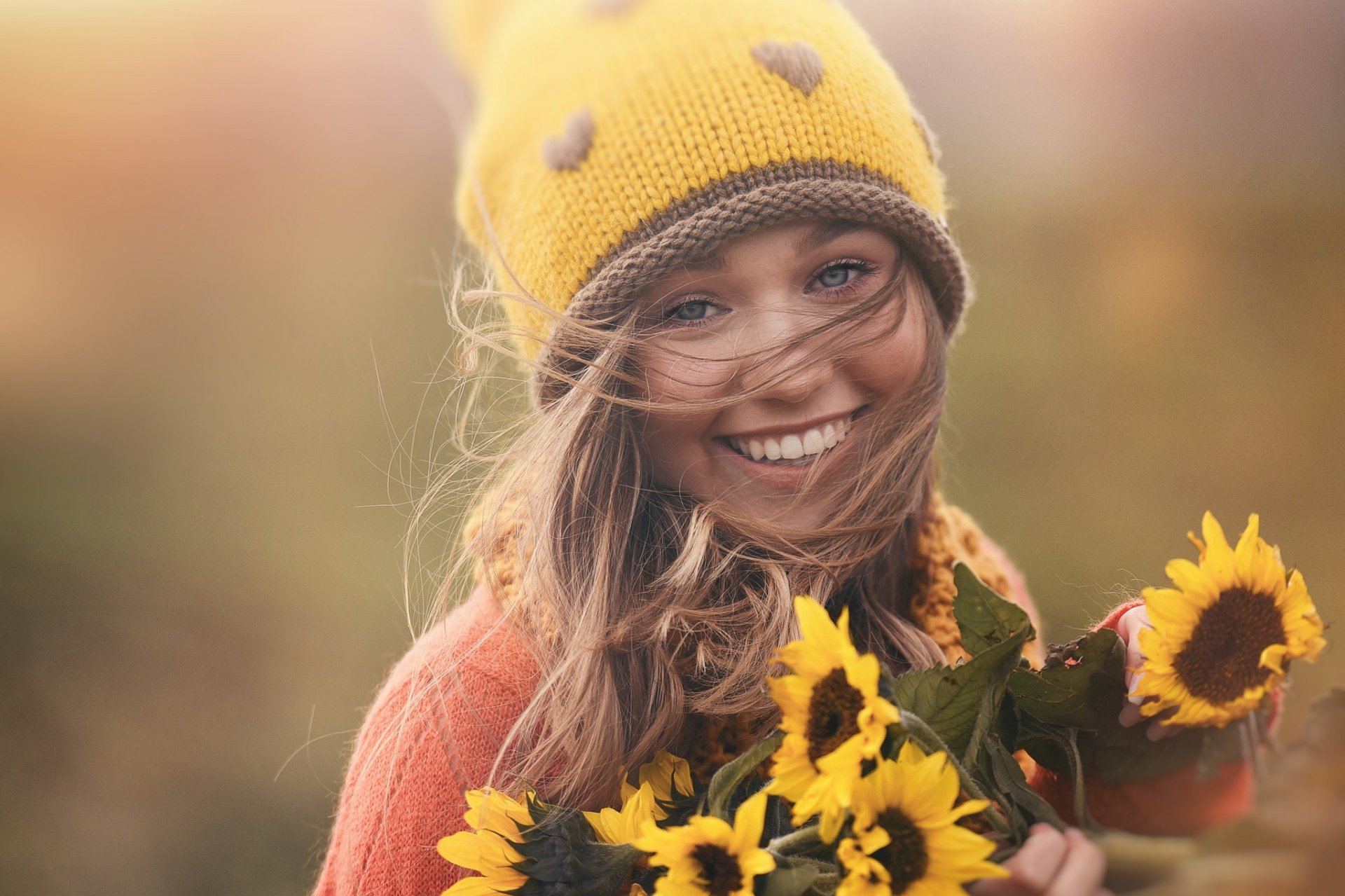 A smiling brunette woman with blue eyes wearing a yellow hat holds bright sunflowers, captured in an HD desktop wallpaper background.