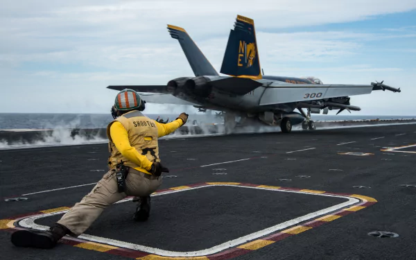 A McDonnell Douglas F/A-18 Hornet warplane taking off from an aircraft carrier, with a deck crew member guiding the jet fighter. Military HD desktop wallpaper.