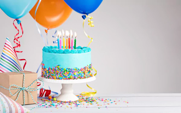 A festive HD birthday desktop wallpaper featuring a bright, colorful cake with candles, balloons, confetti, and a wrapped gift on a white table.