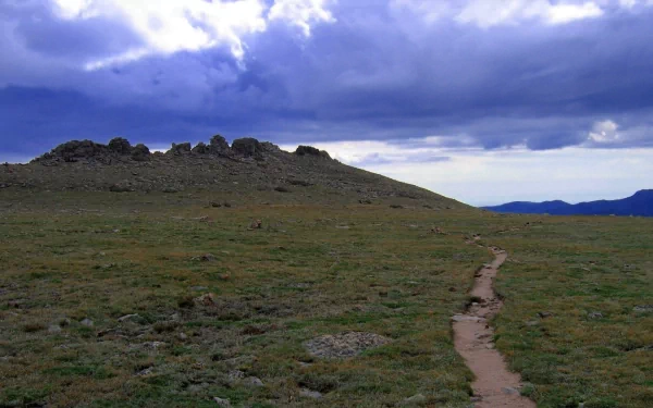 A dirt path winds through a grassy hillside with stone formations under a cloudy sky, capturing a serene mountain landscape in high-definition.