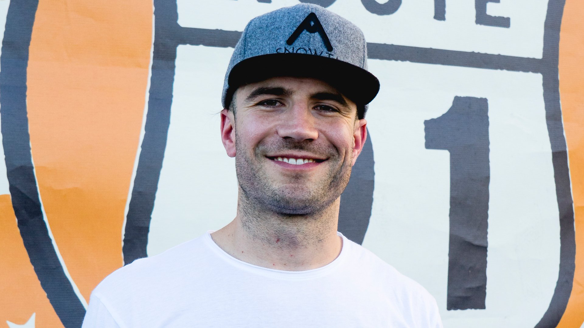 HD desktop wallpaper featuring a smiling man wearing a cap, set against a backdrop with graphic elements.