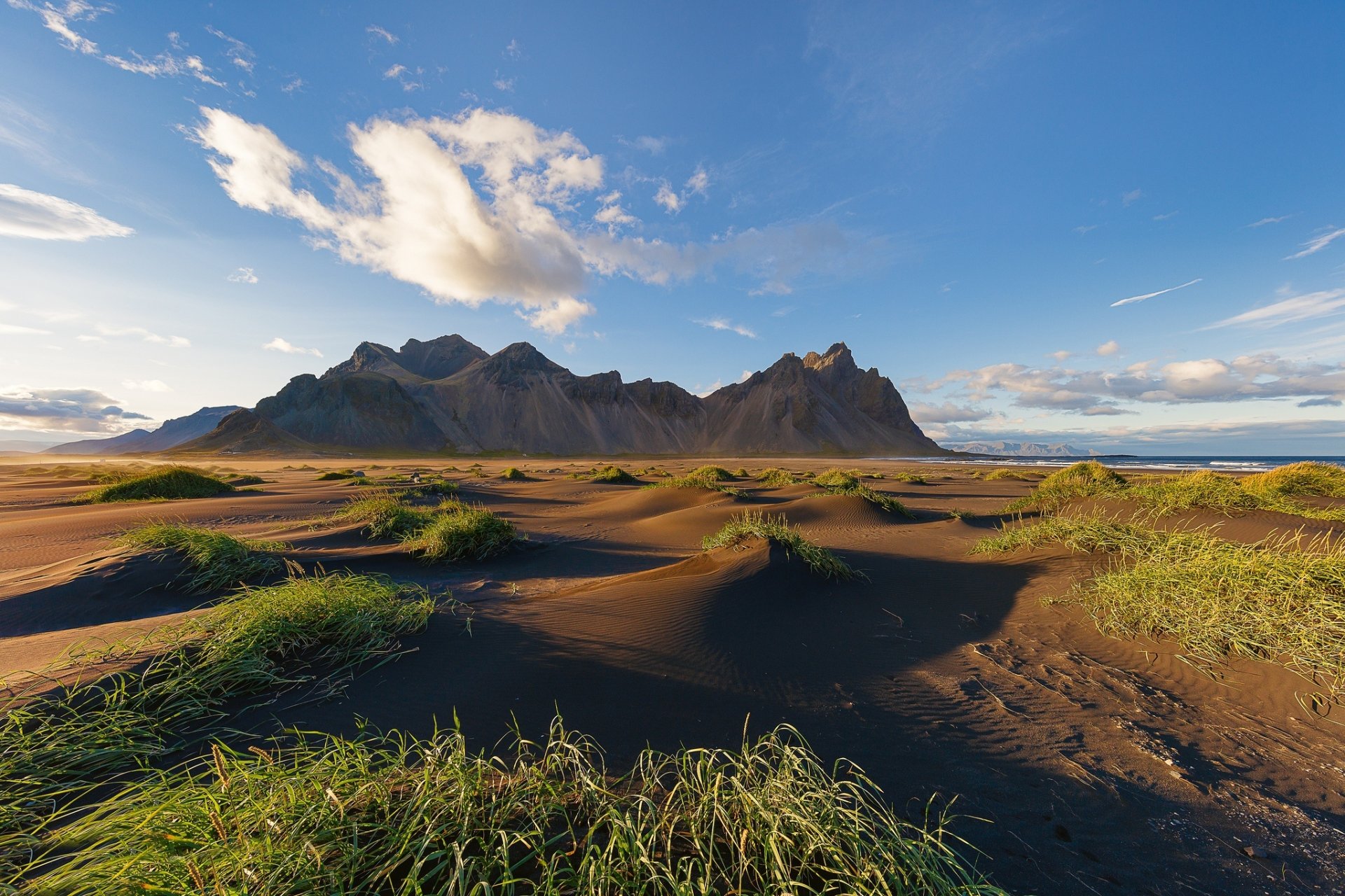 A stunning HD desktop wallpaper showcasing Vestrahorn in Iceland, featuring a dramatic landscape with sand dunes, scattered grass tufts, and a clear sky.