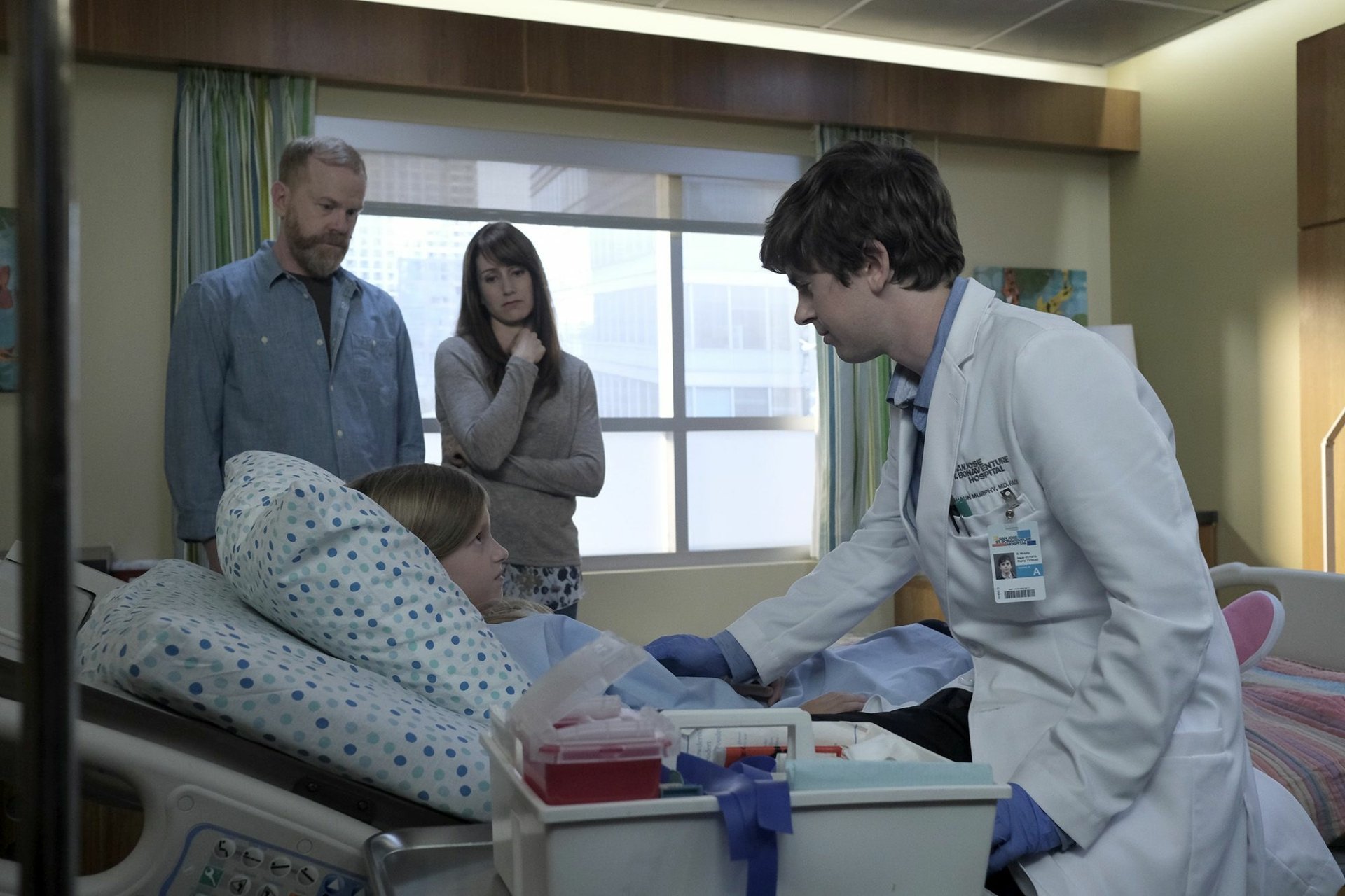 HD wallpaper of 'The Good Doctor' with a doctor treating a patient in a hospital room, as two individuals look on.