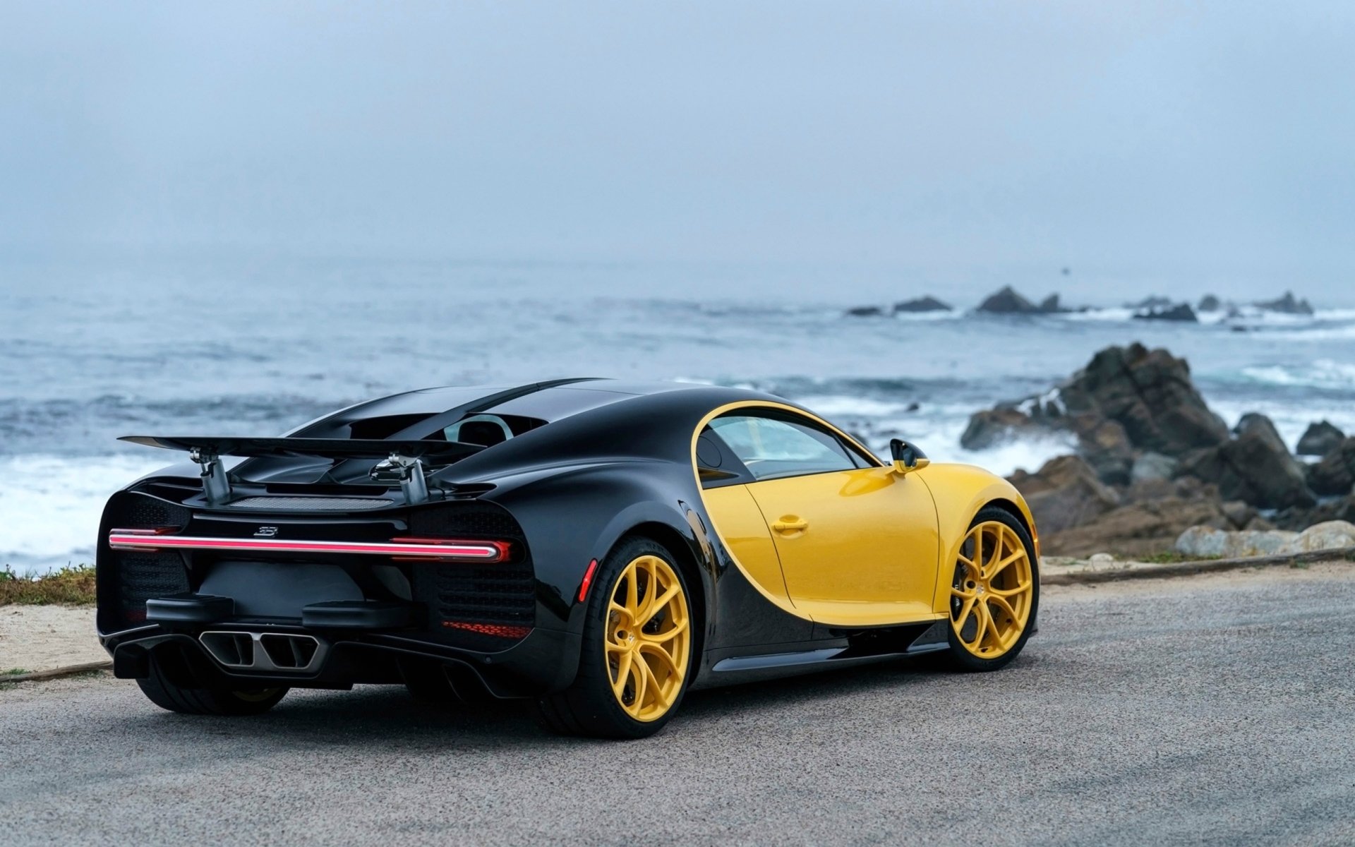 HD PC desktop wallpaper featuring a sleek black and yellow Bugatti Chiron parked by a rocky coastline under a cloudy sky.