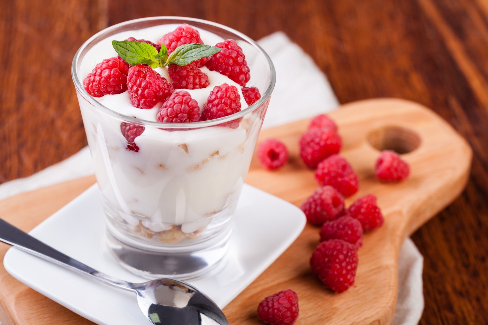 Close-up of a glass filled with yogurt topped with fresh raspberries and a mint leaf, surrounded by loose raspberries on a wooden board, captured in 4K Ultra HD.