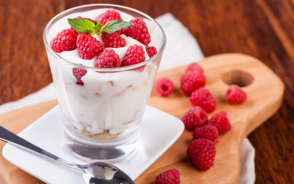 Close-up of a glass filled with yogurt topped with fresh raspberries and a mint leaf, surrounded by loose raspberries on a wooden board, captured in 4K Ultra HD.