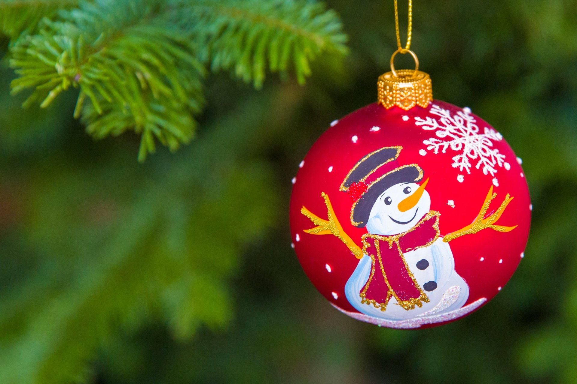A red Christmas bauble with a snowman design hangs from a pine branch, captured with a festive holiday depth of field in an HD desktop wallpaper.
