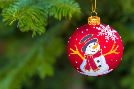 A red Christmas bauble with a snowman design hangs from a pine branch, captured with a festive holiday depth of field in an HD desktop wallpaper.
