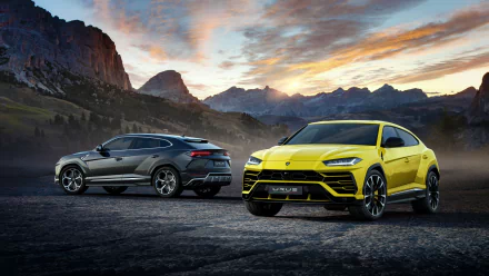 HD desktop wallpaper featuring a stunning Lamborghini Urus SUV duo: a sleek silver car and a vibrant yellow car, set against a dramatic mountain landscape at sunset.