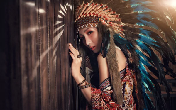 A high-resolution 4K image of an Asian woman model with brown eyes wearing a Native American feathered headdress, leaning against a wooden surface.
