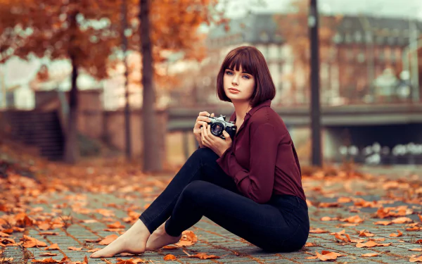 A woman with short hair sits barefoot on a leaf-covered path in fall, holding a camera, captured with depth of field in an HD desktop wallpaper by Marie Grippon.