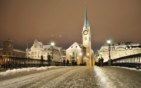 Snow-covered street in Zurich at night, illuminated by warm lights, featuring a historic religious church against a winter sky in Switzerland. 4K Ultra HD wallpaper.