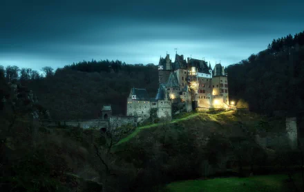 Eltz Castle in Germany illuminated at dusk, nestled among dark forested hills, displayed as a high-definition PC desktop wallpaper.