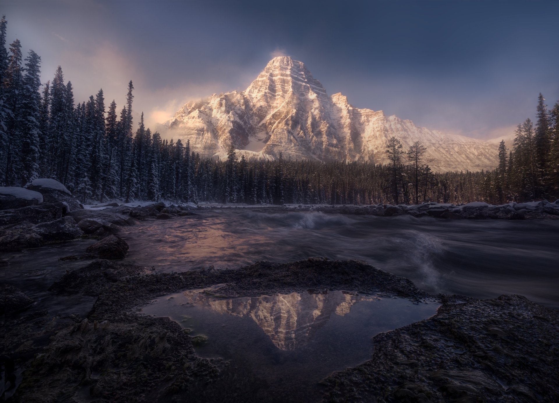Snow-covered mountain peak reflecting in a calm river, surrounded by a dense winter forest. The image captures the serene beauty of nature and is a HD desktop wallpaper and background.