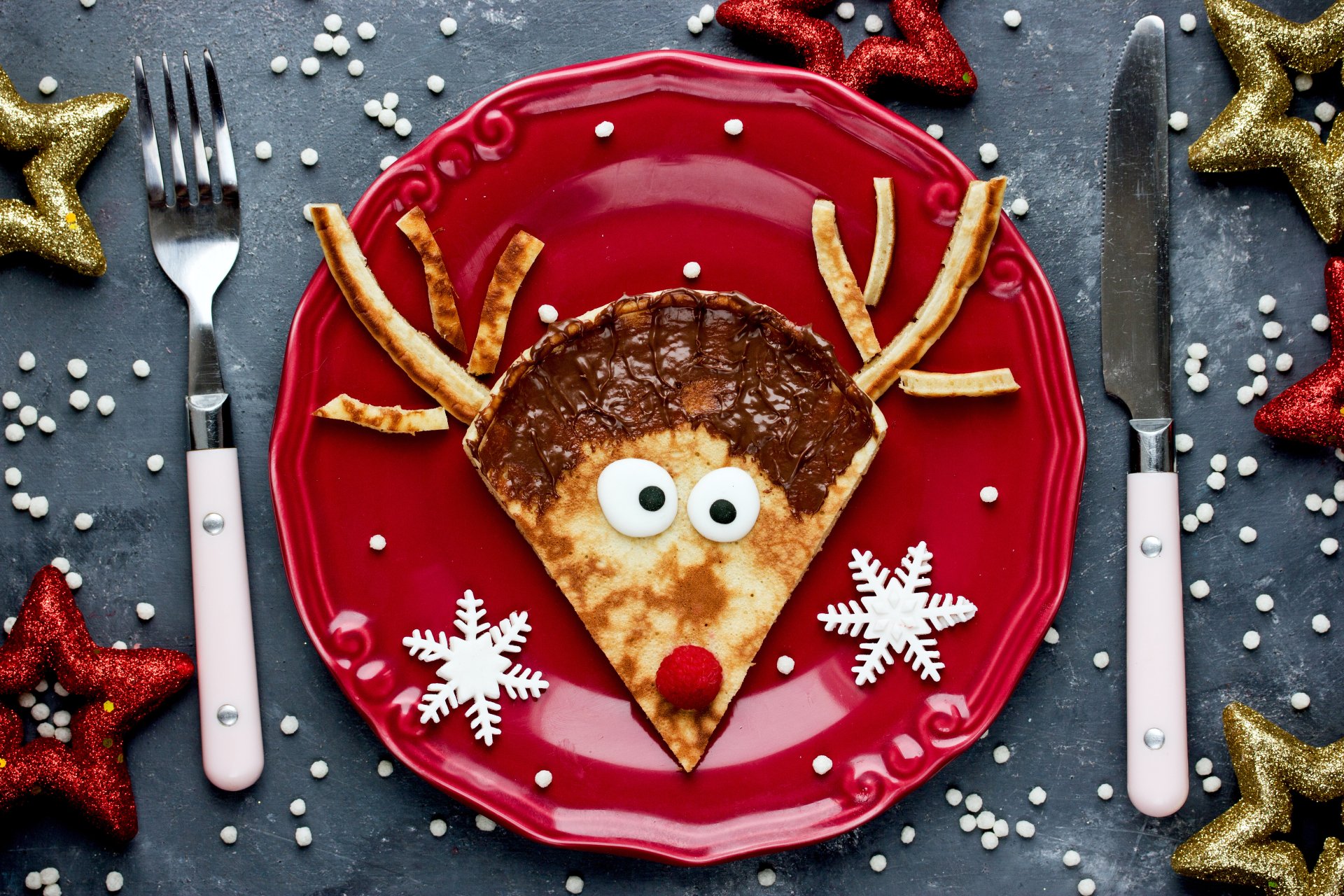 A humorous still life 4K Ultra HD wallpaper featuring a crêpe decorated as a reindeer on a red plate with snowflake-shaped accents and festive gold and red stars.
