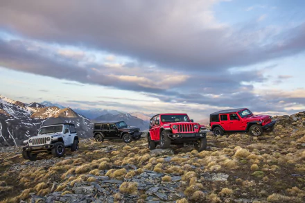 Four Jeep Wrangler SUVs, including white, black, and red models, are parked on rocky terrain with mountains and a cloudy sky in the background.