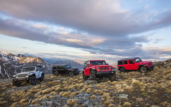 Four Jeep Wrangler SUVs, including white, black, and red models, are parked on rocky terrain with mountains and a cloudy sky in the background.
