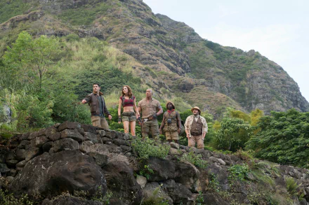 HD desktop wallpaper featuring Kevin Hart, Karen Gillan, Jack Black, and Dwayne Johnson in a scene from the movie Jumanji: Welcome to the Jungle. The characters stand on a rocky terrain with lush hills in the background.