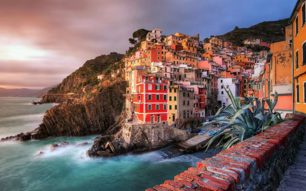Colorful houses of Riomaggiore, Italy, perched on a rugged coastal cliff under a moody sky, showcasing a vibrant man-made cityscape by the sea.
