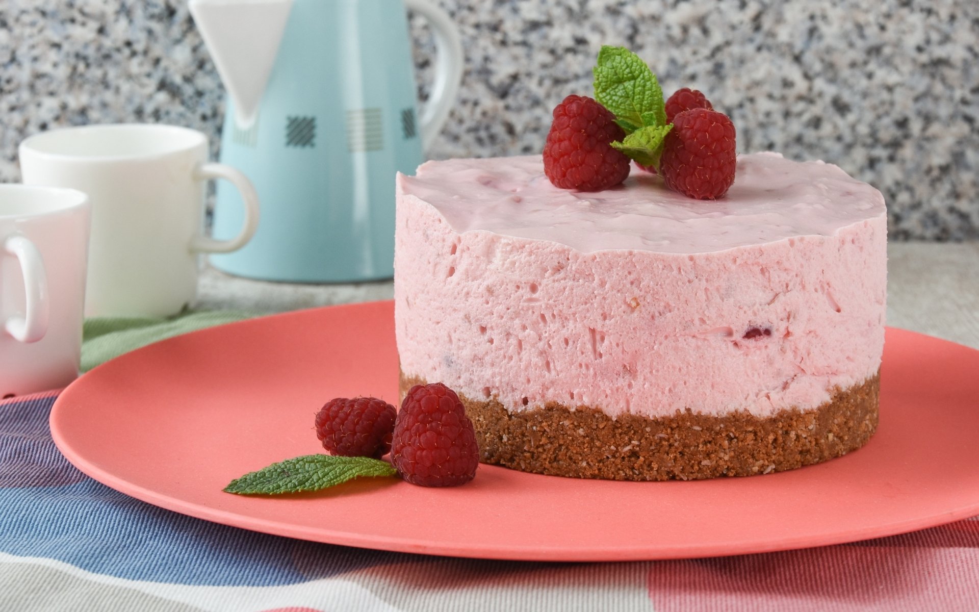 HD desktop wallpaper featuring a pink raspberry mousse cake with a crumb crust, garnished with fresh raspberries and mint on a coral plate in a kitchen setting.