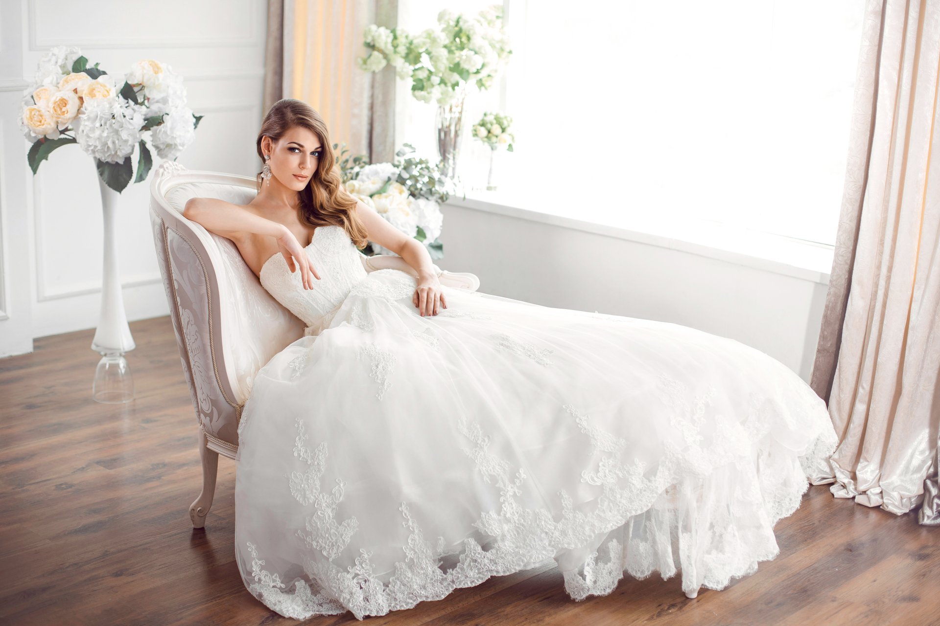 A brunette bride in a white wedding dress and earrings lounges gracefully on a chair by a sunlit window, captured in an HD desktop wallpaper.