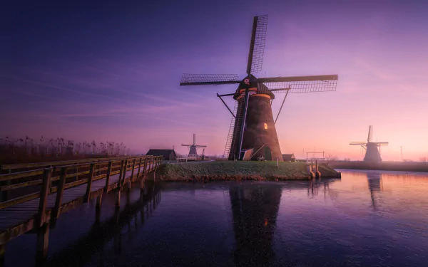 HD desktop wallpaper featuring a serene sunrise with a clear sky reflecting off the water. Traditional windmills and a wooden pathway create a picturesque scene, blending man-made structures with natural beauty.