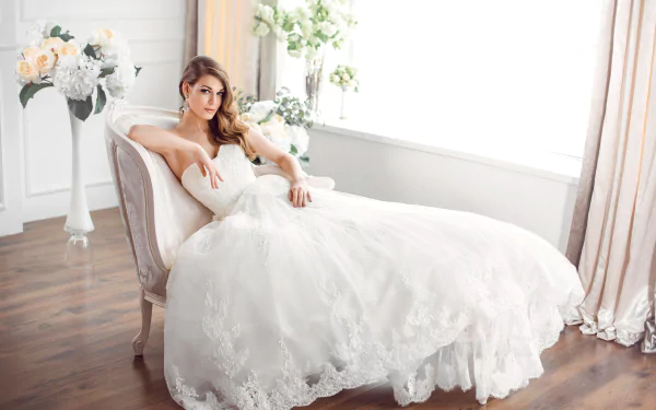 A brunette bride in a white wedding dress and earrings lounges gracefully on a chair by a sunlit window, captured in an HD desktop wallpaper.