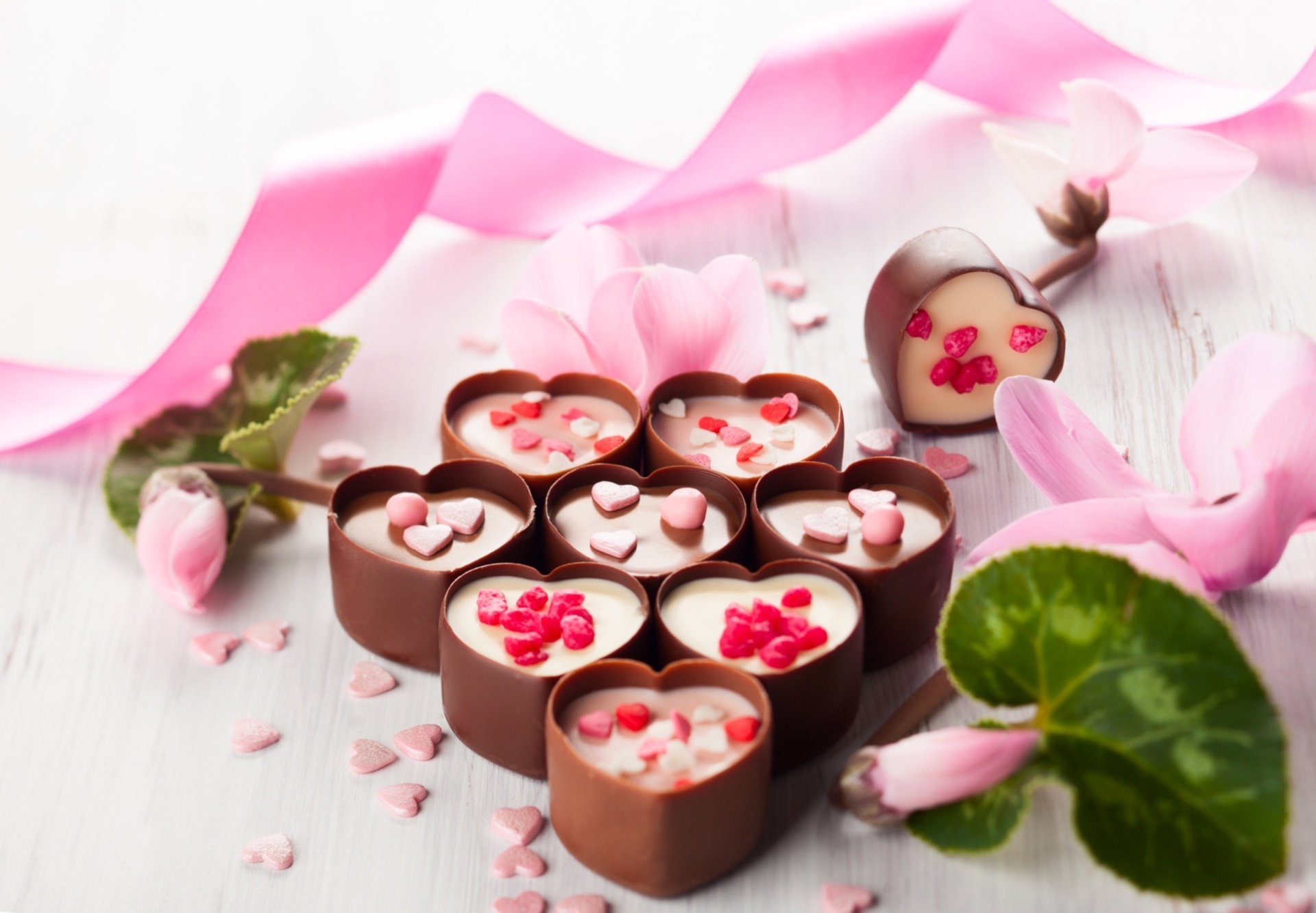 HD desktop wallpaper featuring heart-shaped chocolates decorated with pink and red candy hearts, surrounded by pink ribbon and flowers, evoking love and sweetness.